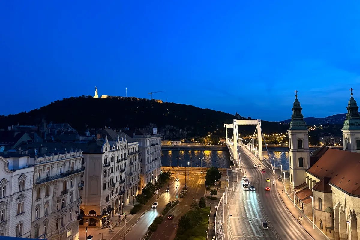 The Szechenyi bridge crossing the Danube at nighttime, buildings on either side, and a hill in the distance.