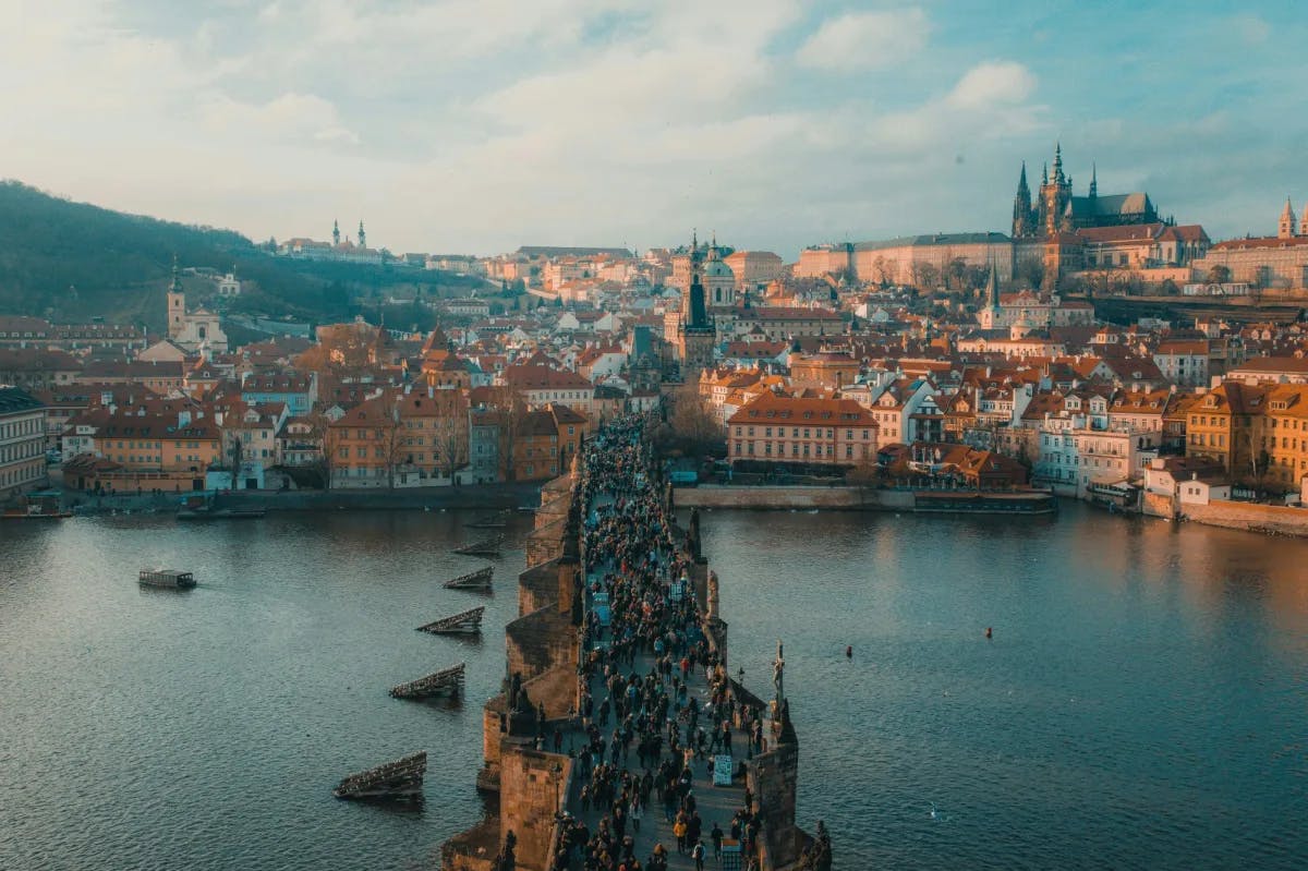 An aerial view of people walking down a stone bridge in Prague atop of a river with the city scape in the background. 