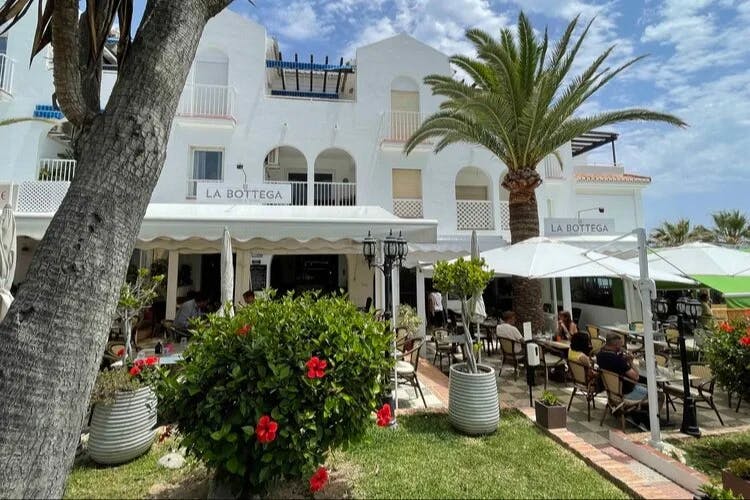 A large beautiful white building with outdoor patio seating, palm trees, red flowers and umbrellas for shade at La Bottega Food & Wine an Italian laid-back restaurant with a terrace & sea views.