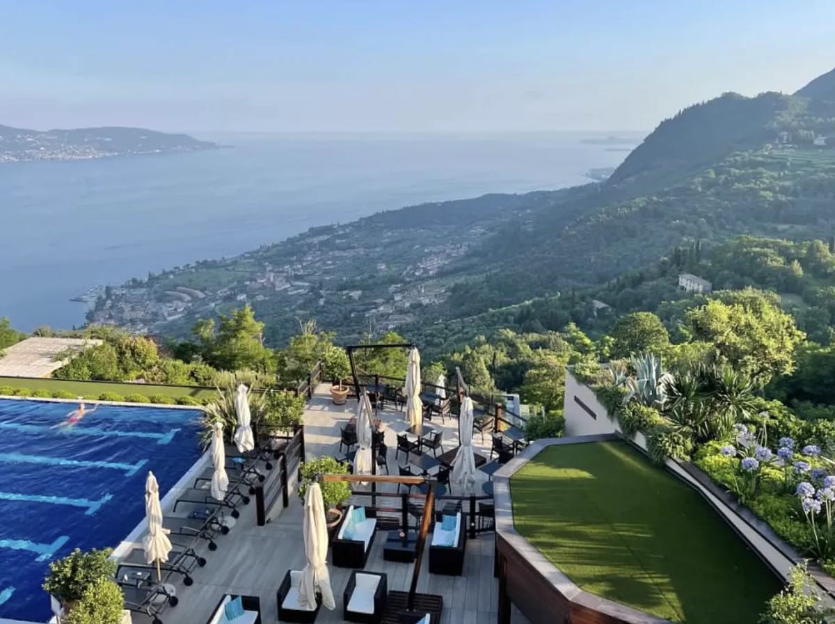 An aerial view of a hotel patio with a swimming pool, lounge seating, grassy lawn and stunning view of the lake and mountains. 