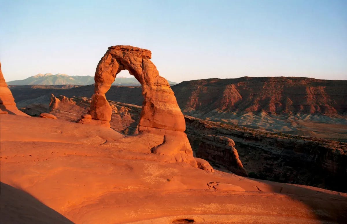 Brown rock arch with a view of the canyon behind it at sunset.