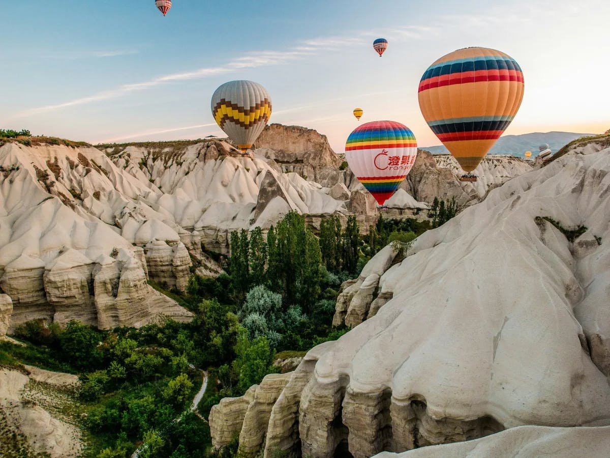 Colorful hot air balloons soar above a rugged landscape dotted with rock formations and verdant patches.