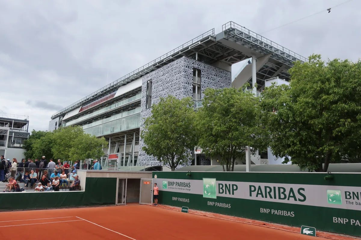 A large grey building towering near a red tennis court and lots of people sitting in the audience section. 