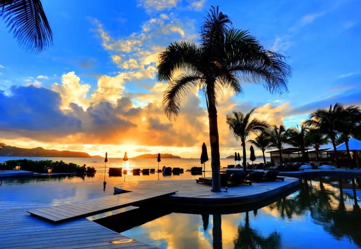 pool flanked by a chic wooden deck, set against a stunning sunset and palm trees