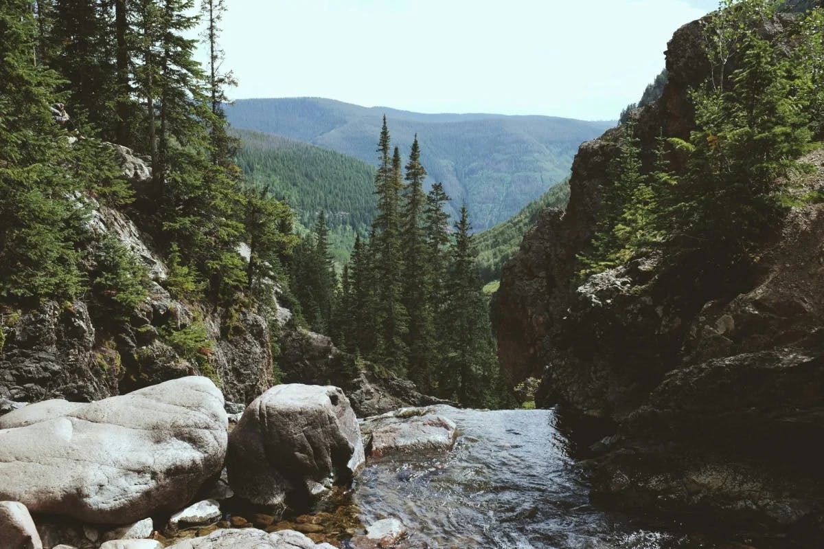 stream and boulders at the top of a trail with forests and mountains in the background