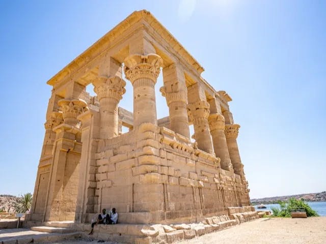 A stone temple in the desert next to a river with people sitting on it