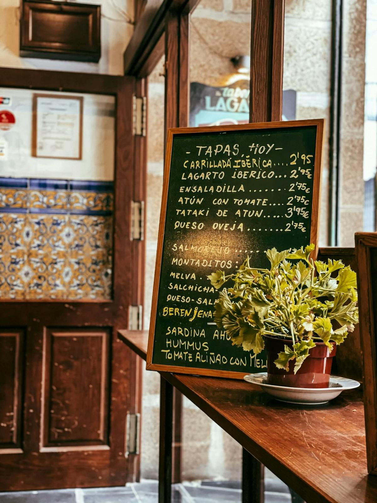 A chalkboard menu displayed on a wooden surface inside of a bright hotel, with a small, potted plant in front of it. 