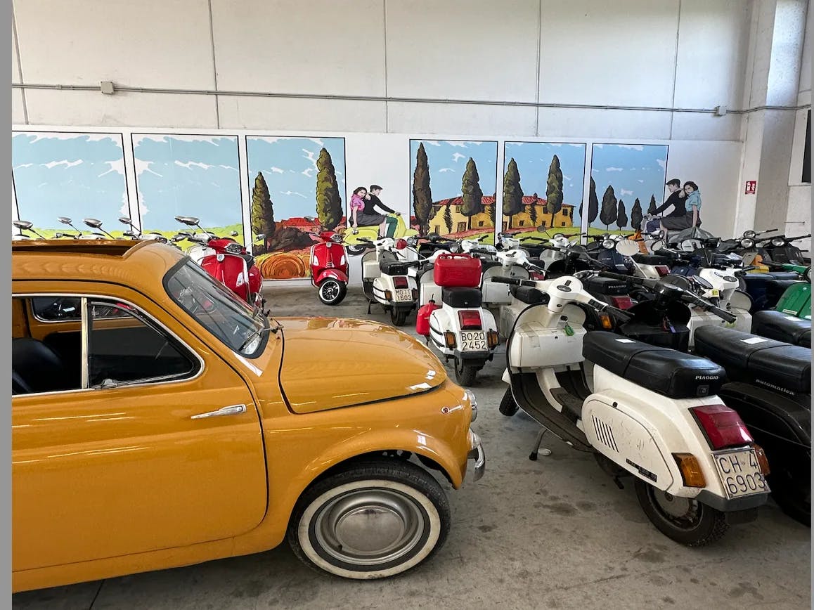 A collection of scooters and a classic yellow car are showcased in a room adorned with outdoor scene paintings.