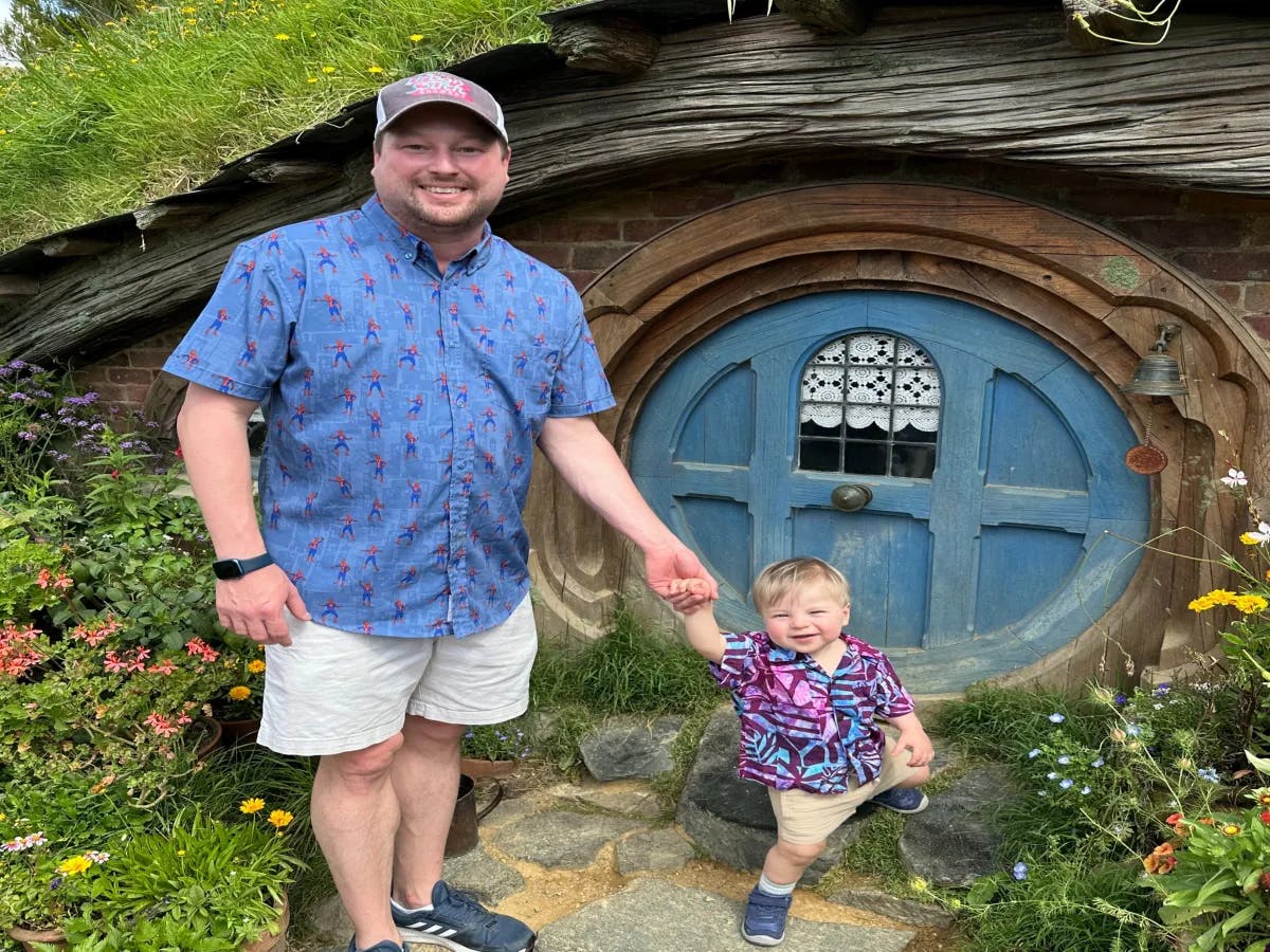 An adult and a child stand in front of a round door of a Hobbiton house during the daytime.