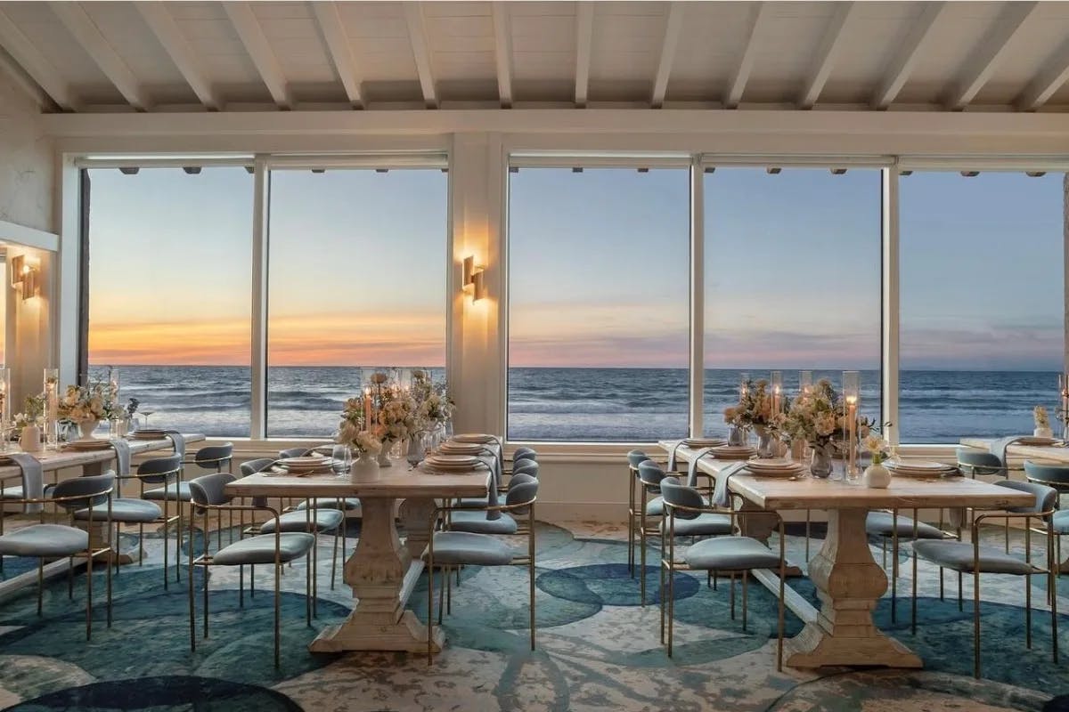 The Marine Room, a restaurant with white walls, blue carpet and large floor-to-ceiling windows looking out at the ocean and sunset.