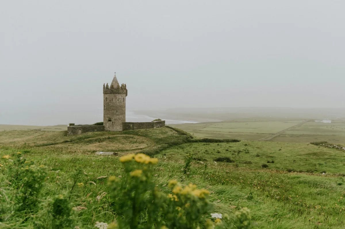 A castle in a green field with the sea in the distance on a cloudy day.