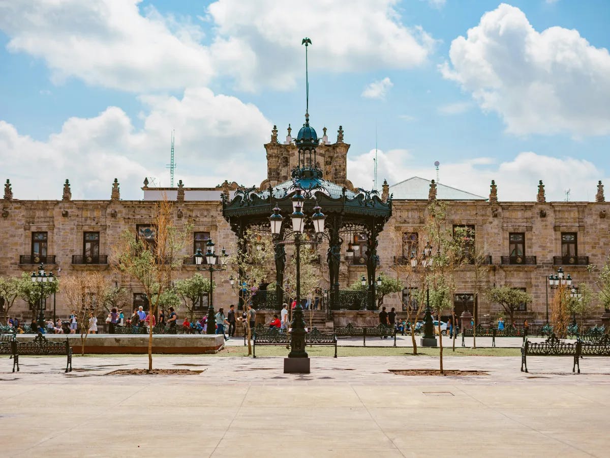 The image shows a bustling public square with a decorative gazebo at the center, surrounded by trees and benches, with a large building in the background under a blue sky with clouds.