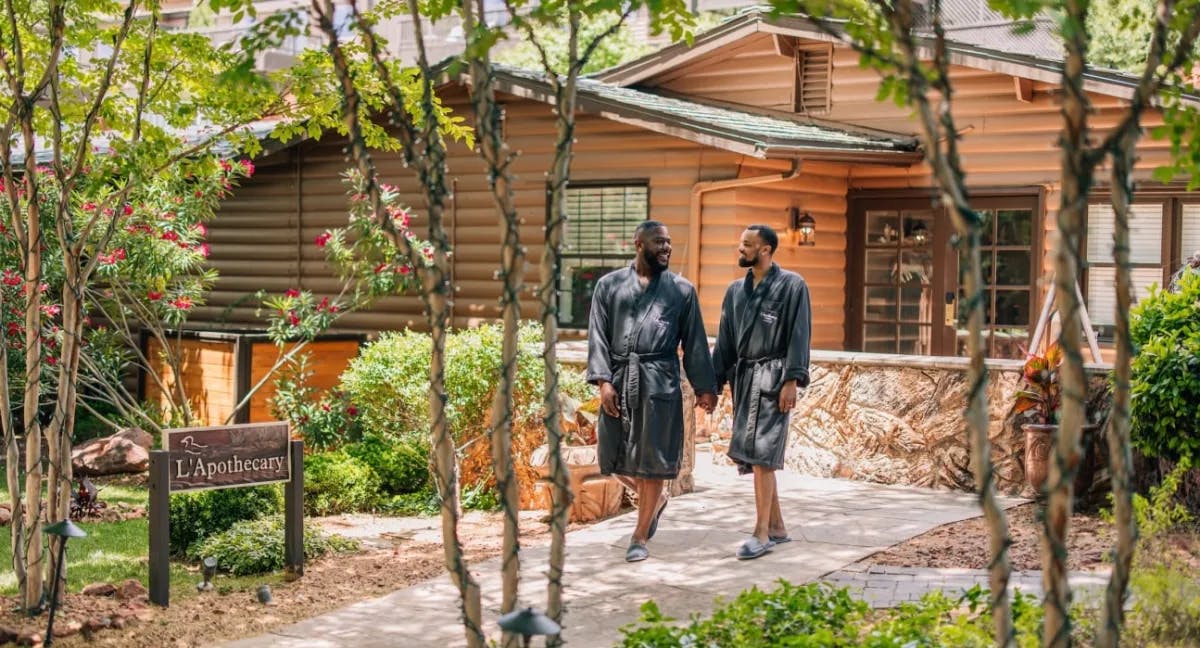 Two men walking outside of a spa wearing robes and holding hands. There are trees, a wooden sign and a log house in the surrounding areas. 