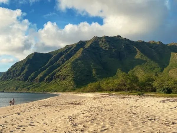 The image shows two individuals walking along a sandy beach towards the ocean, with a lush green mountain range in the background under a blue sky.