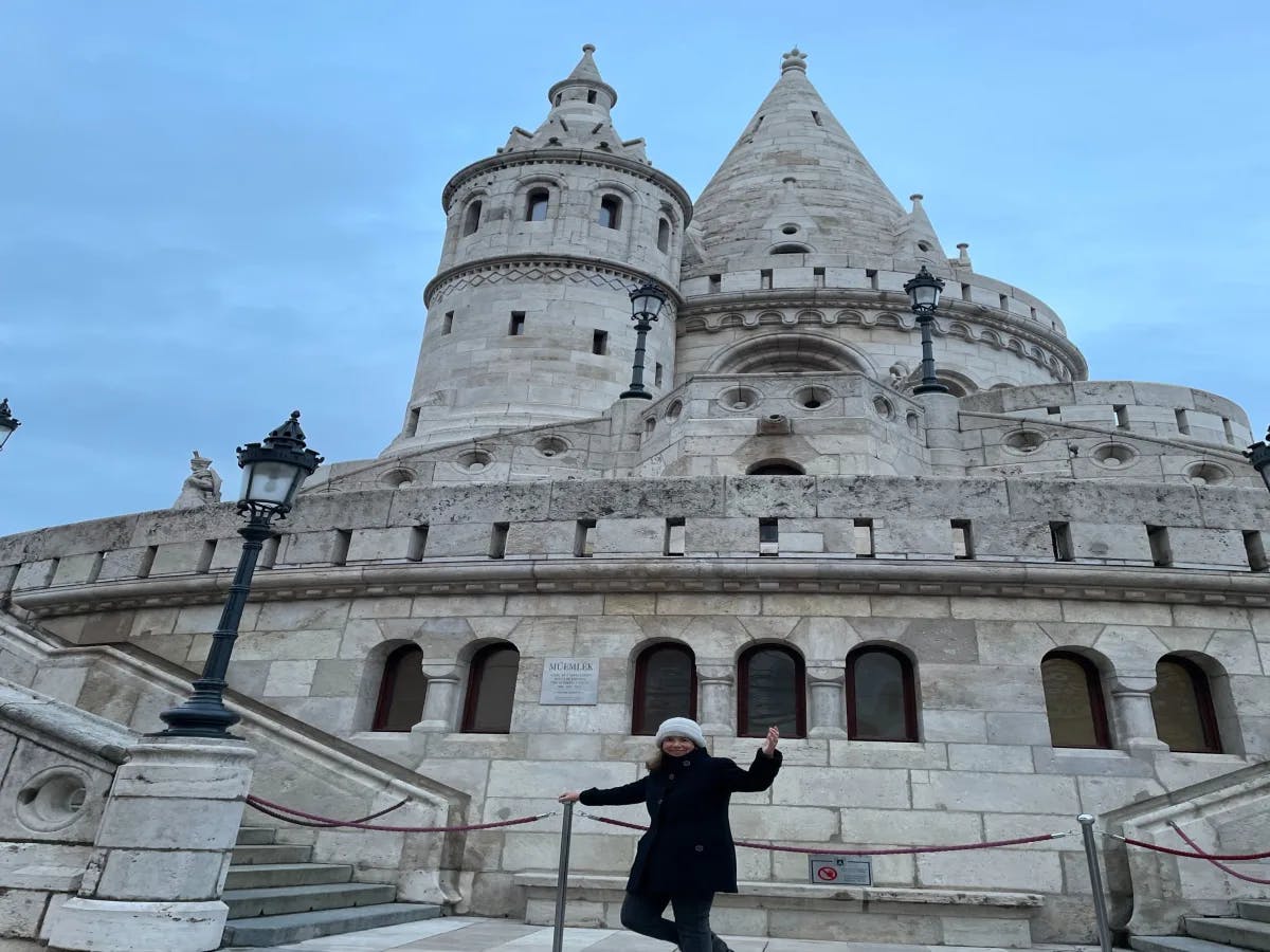 A person in black winter clothes with arms outstretched, standing in front of an ornate, historic stone building with architectural details