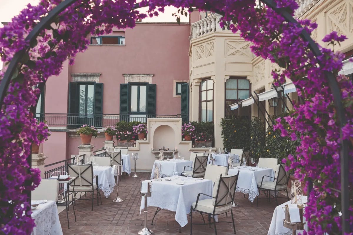 terrace with dining tables with an archway of purple flowers and a pink hotel in the background