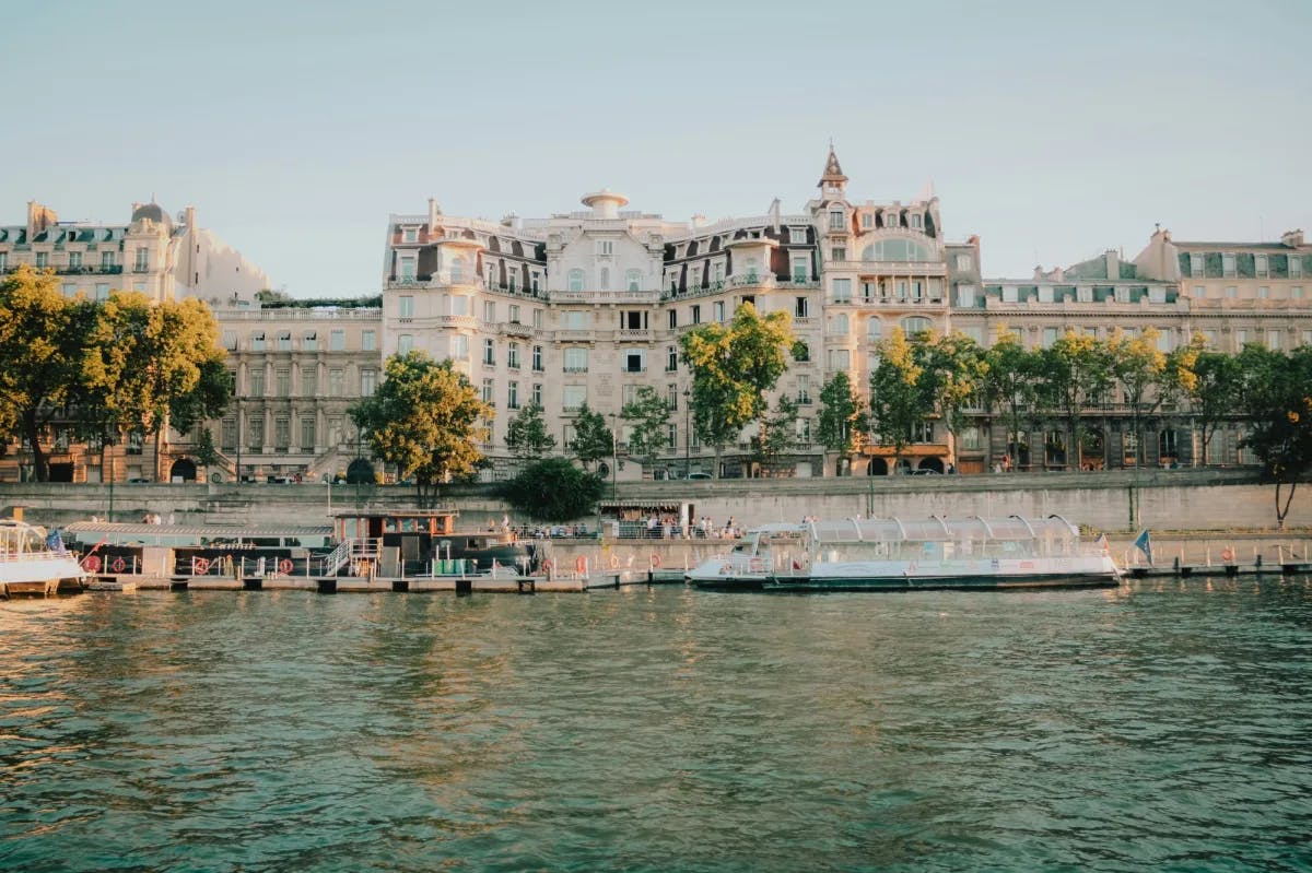 An image that depicts an extravagant Parisian building with a river in the forefront view. There is a boat, trees and people walking along it. 