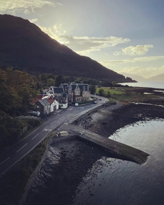 An aerial view of the Ballachulish Hotel surrounded by water, a mountain and green trees on a cloudy day.
