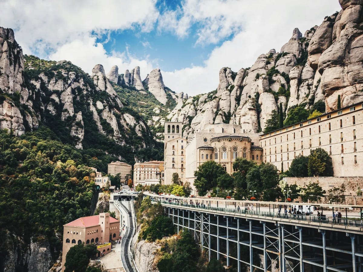 A majestic mountainous landscape with a monastery built into the cliffs, connected by a bridge to a road, under a partly cloudy sky.