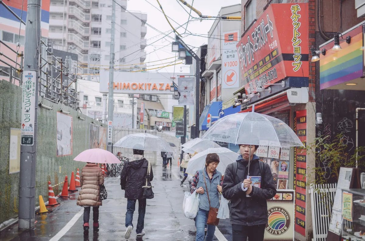 People walking down a street in front of shops in Shimokitazawa holding umbrellas during daytime.