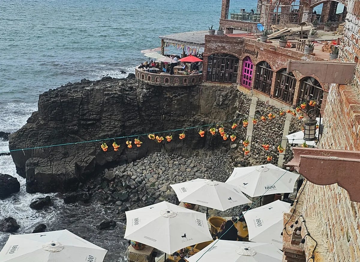 The image captures a seaside dining area with white umbrellas, framed by rocky cliffs and the ocean.