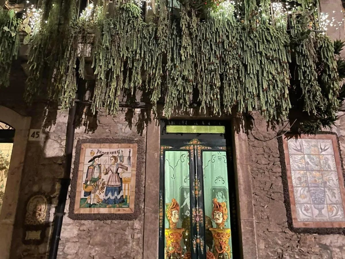 The image shows a richly decorated facade of a building with hanging plants, religious artwork, and ornate doors.