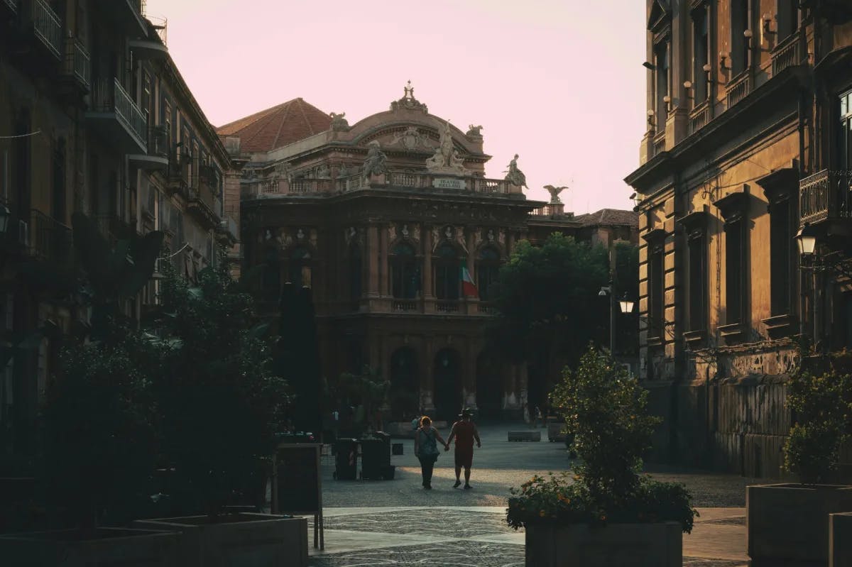 Two people walking down a street surrounded by tall historical buildings with ornate architecture at sunset.