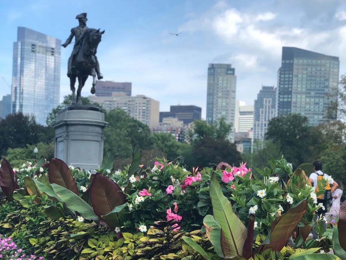 A vibrant garden with various flowers, a statue on a pedestal and skyscrapers in the background under a partly cloudy sky.