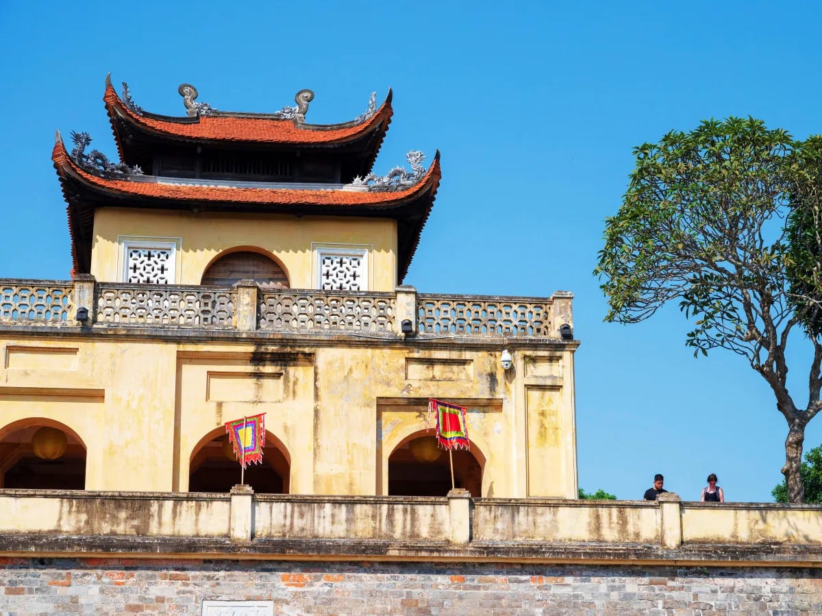 An ancient yellow building with traditional architectural elements under a clear blue sky, flanked by a large tree.