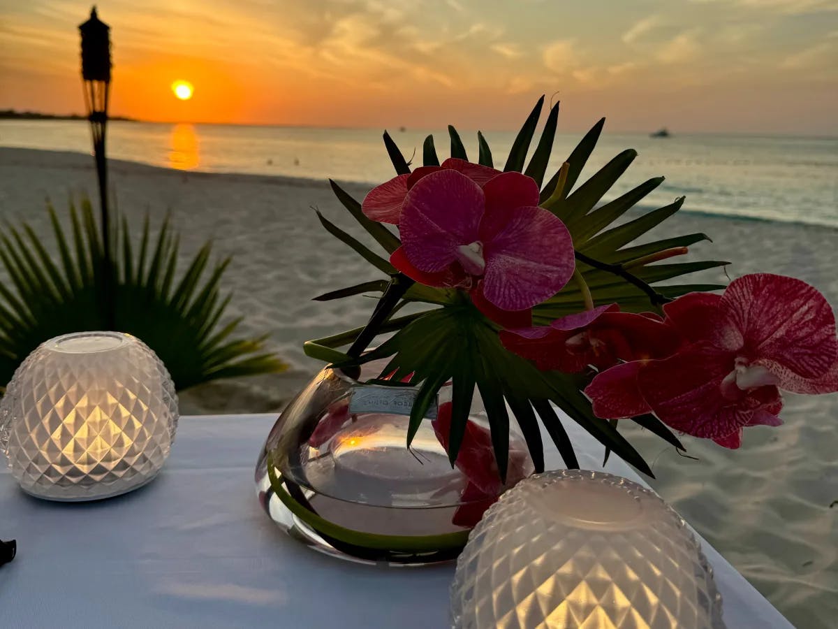 The image presents a tranquil beach sunset with a romantic table setting, complete with flowers and glowing spherical lamps.