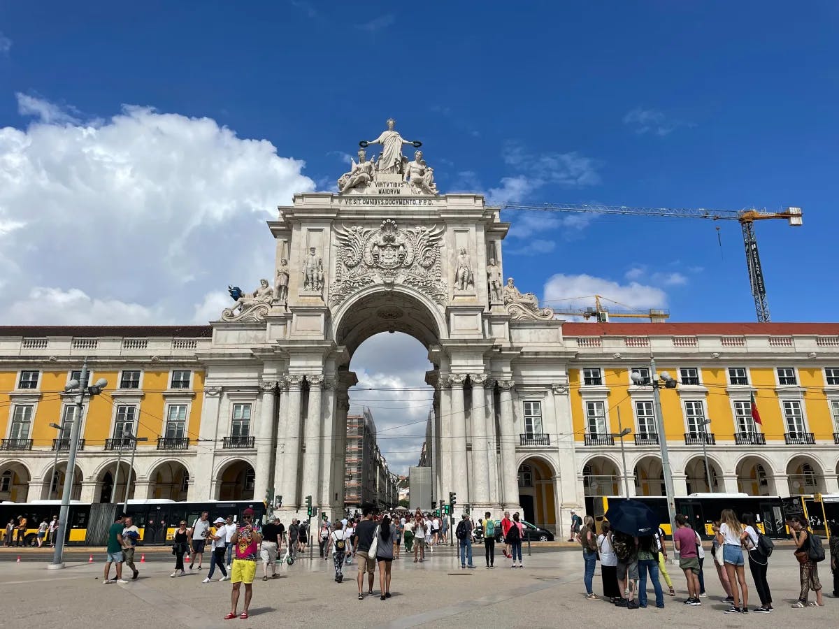 A picture of Praca Do Comercio complete with white and yellow stone detailing with people around during the daytime.