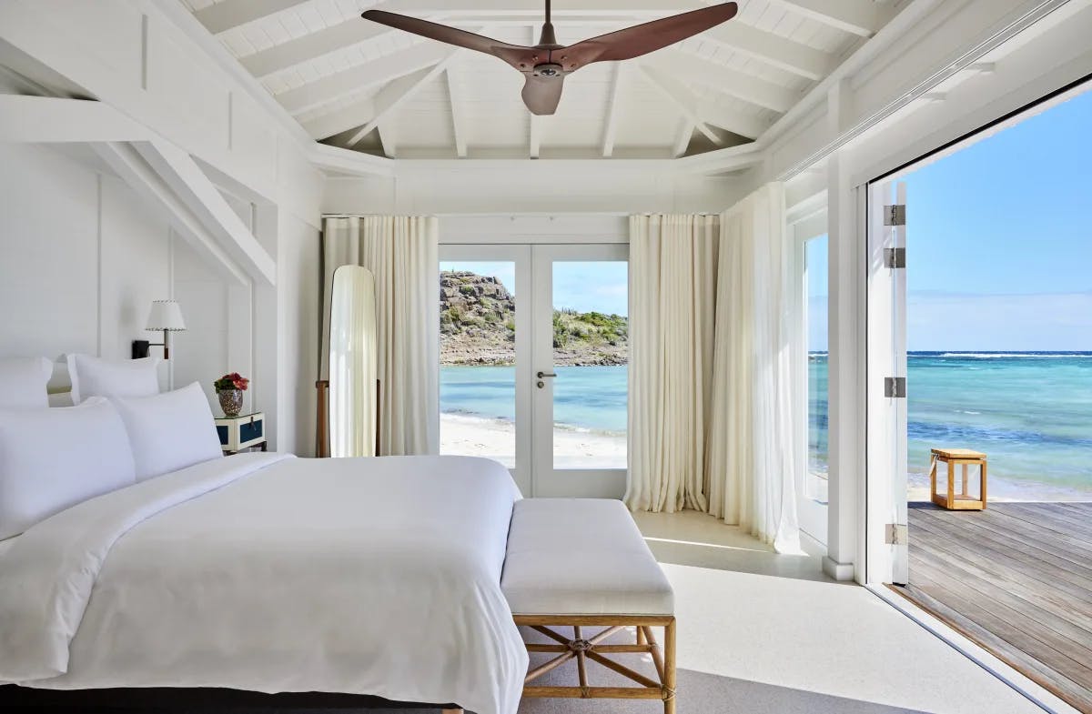 A view of a white bed in a hotel suite next to an open set of doors looking out to the Caribbean sea. 