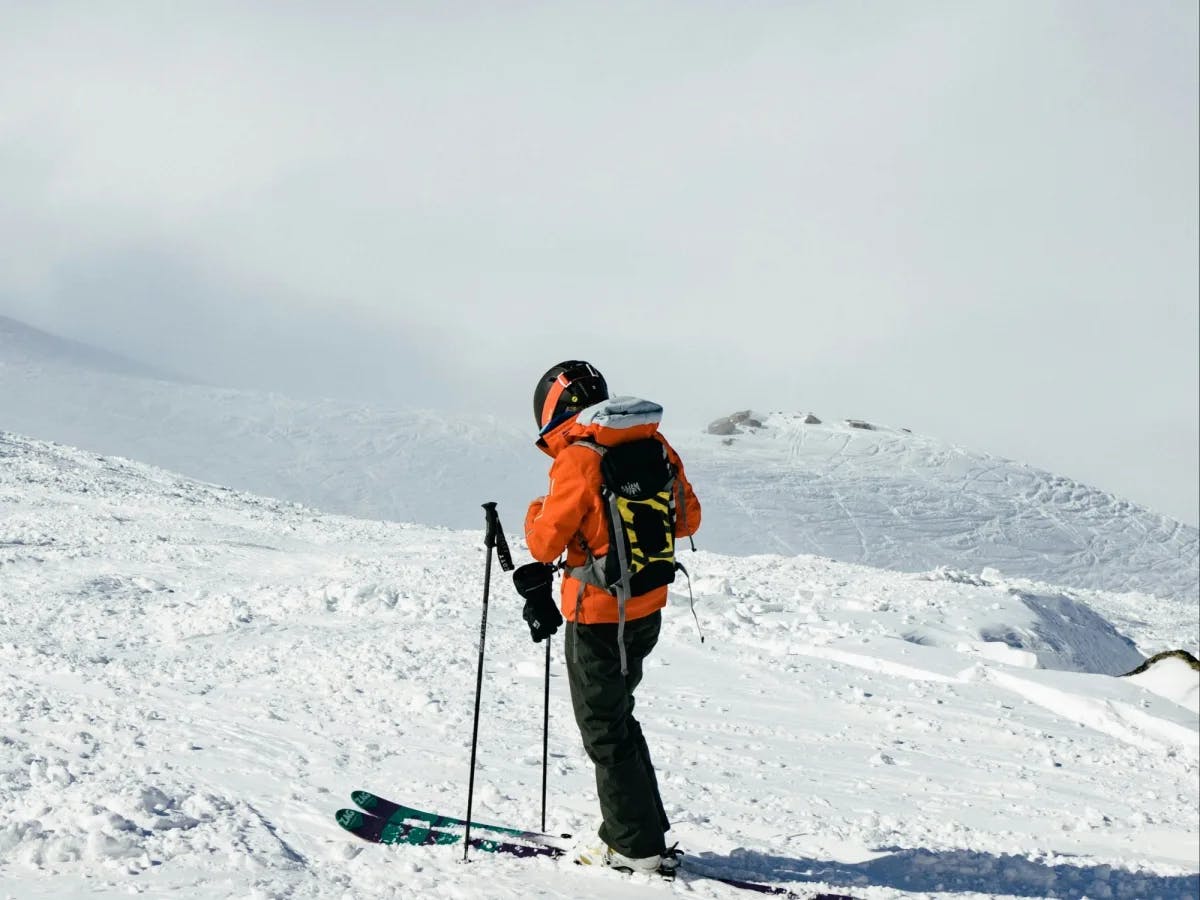 A person wearing an orange jacket, backpack, and skis surrounded by snowy hills and terrain.