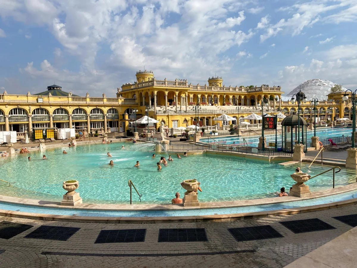 An expansive outdoor thermal bath with classical yellow architecture, filled with bathers under a partly cloudy sky