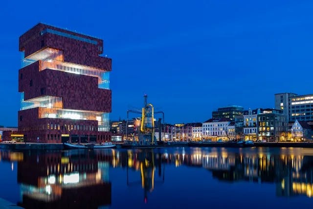 A view of the Museum ann de Stroom (Museum by the River) in Antwerp Belgium, with the river, and other buildings in the background, at night. 