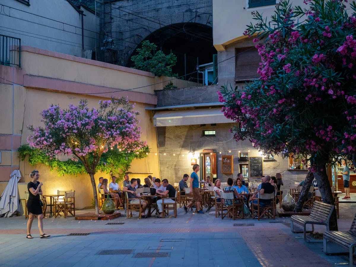 The image features an inviting outdoor dining setting at twilight, with guests enjoying their meals amidst a backdrop of buildings and trees adorned with purple flowers.