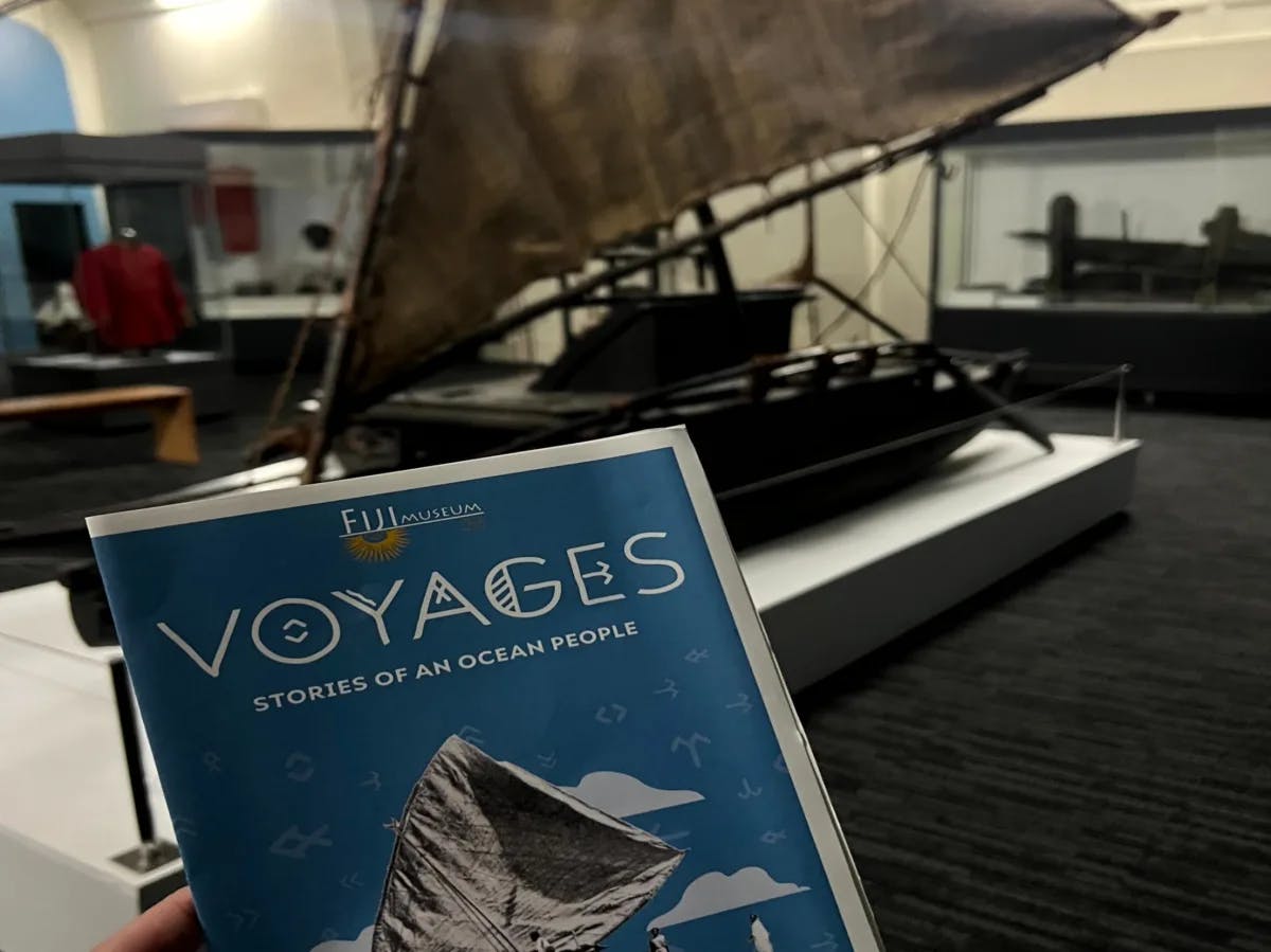 A hand holding a museum brochure titled “VOYAGES” against a backdrop that includes part of a ship’s sail.