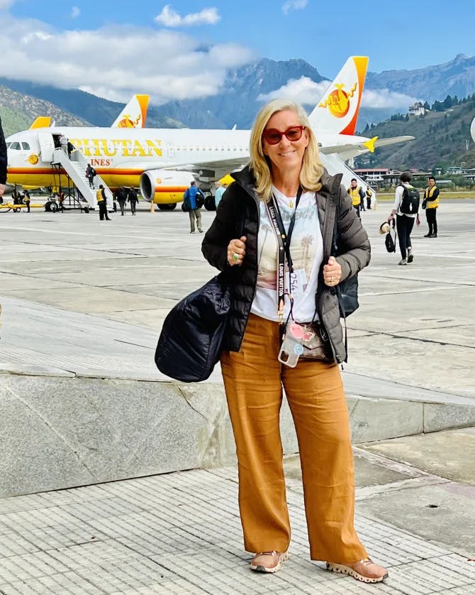 Susanne posing in front of a plane outside while wearing orange pants, a white t-shirt and jacket  