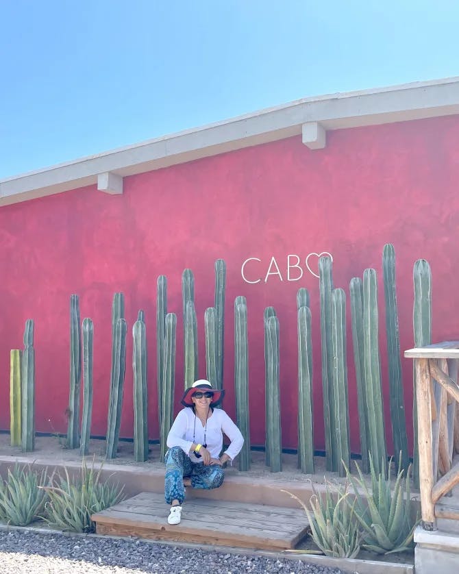 Travel advisor Lisa sitting on a wall in a white shirt and hat in front of a pink wall and cacti with a Cabo sign
