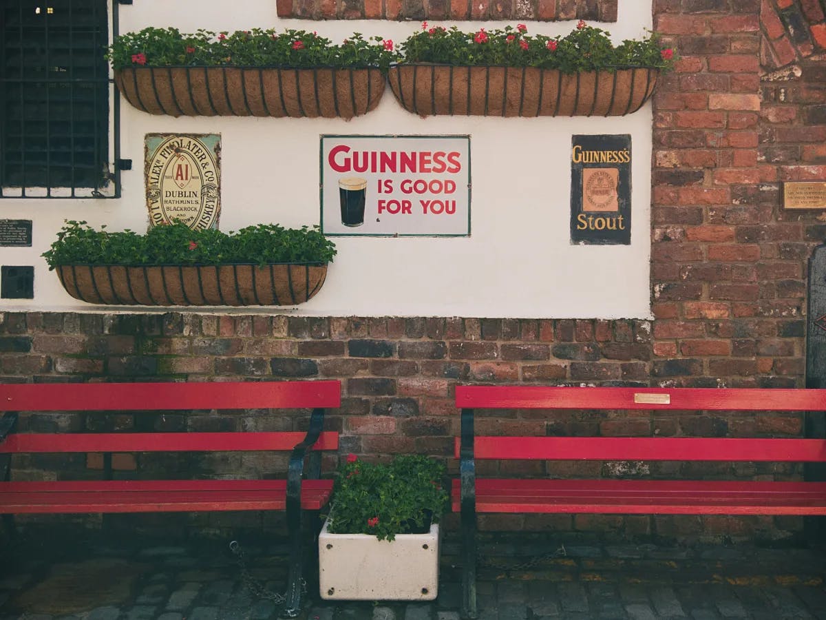 The image shows a red bench with a planter in front, against a brick wall with a sign saying 'Guiness is Good for You' and flower boxes.