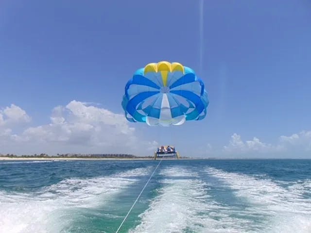 A colorful parasail with blue and yellow stripes glides over the ocean, connected to a boat by a long rope.