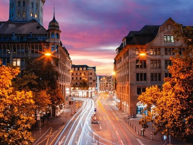 A time lapse photo of a city street in Zurich with trees lit up from the street lamp, stone buildings on each side with lots of windows and a purple sunset in the distance. 