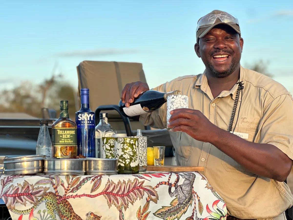 A man pouring wine into a glass, surrounded by an assortment of bottles and glasses on a picnic table, set against a backdrop of lush greenery.