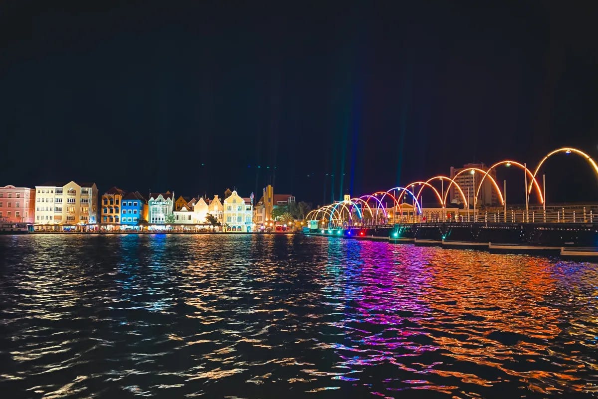 The Queen Emma Bridge, also known as the "Swinging Old Lady," during nighttime lit up with multiple colors reflecting against the water.