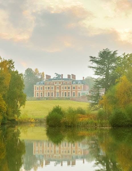 A view of Heckfield Place, a large red building with green roofs, in the distance surrounded by green grass, trees and a reflective body of water. 