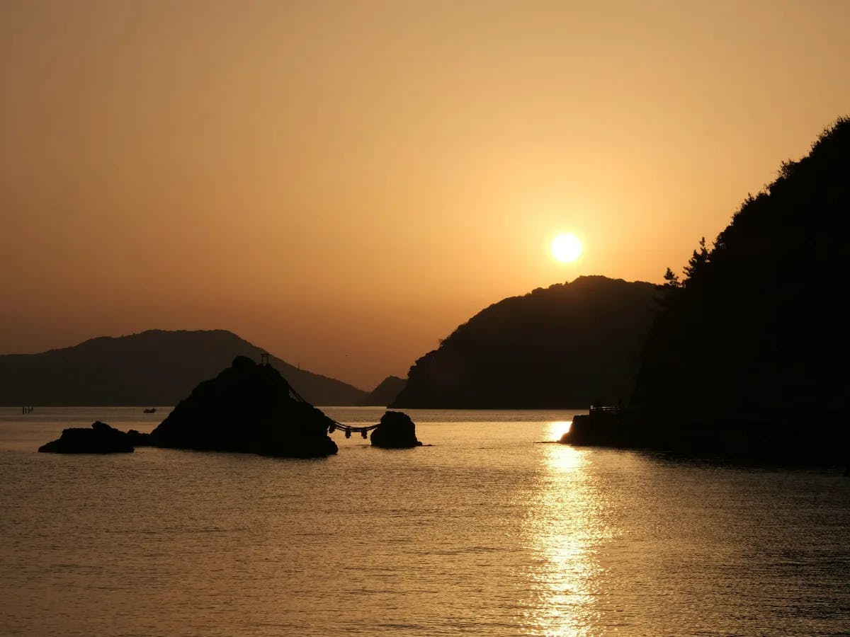 A serene sunset over calm waters with silhouetted hills and landforms in the background.