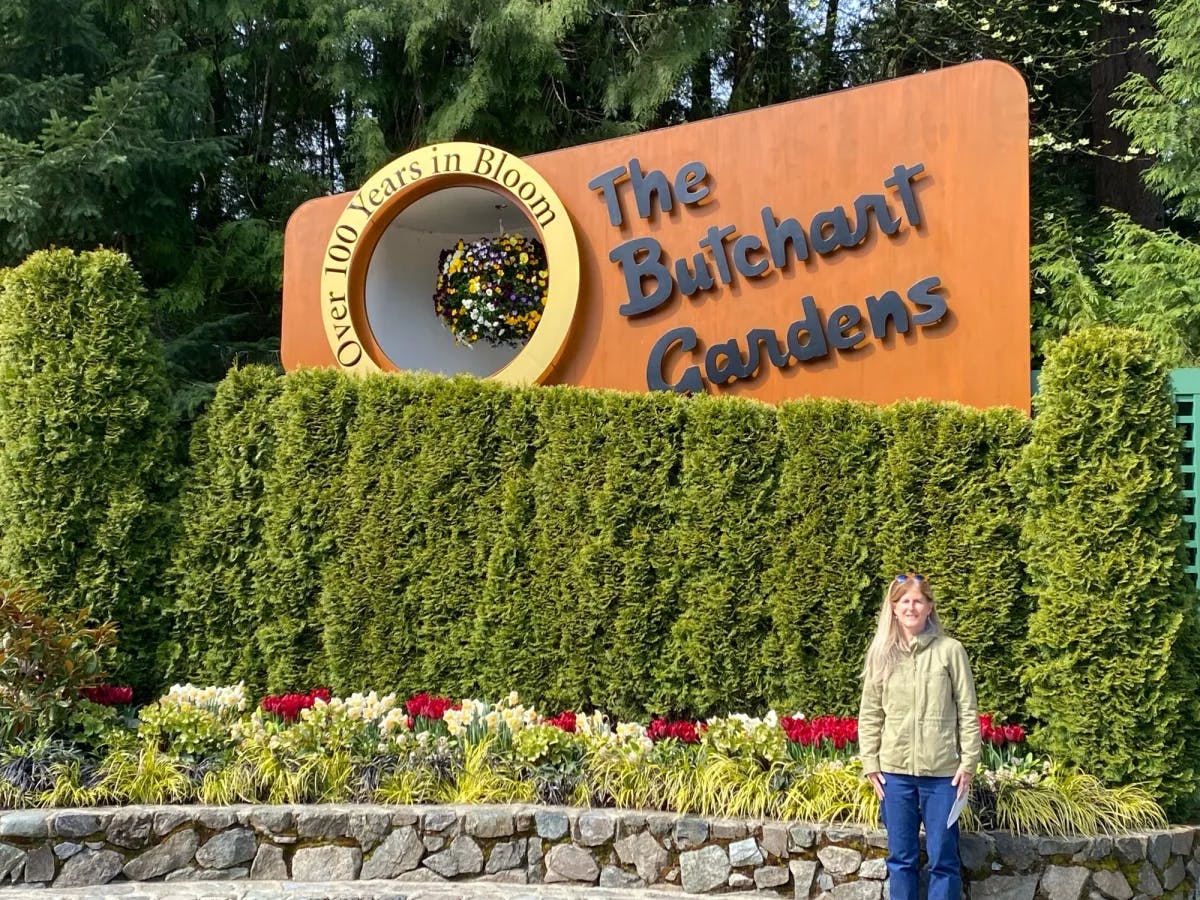 A woman is standing in front of flowers and a big board saying The Butchart Gardens.