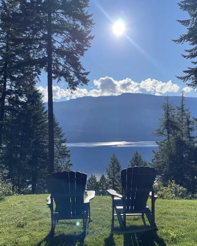 Beautiful view of two chairs on the grass in front of pine trees, lake and mountains