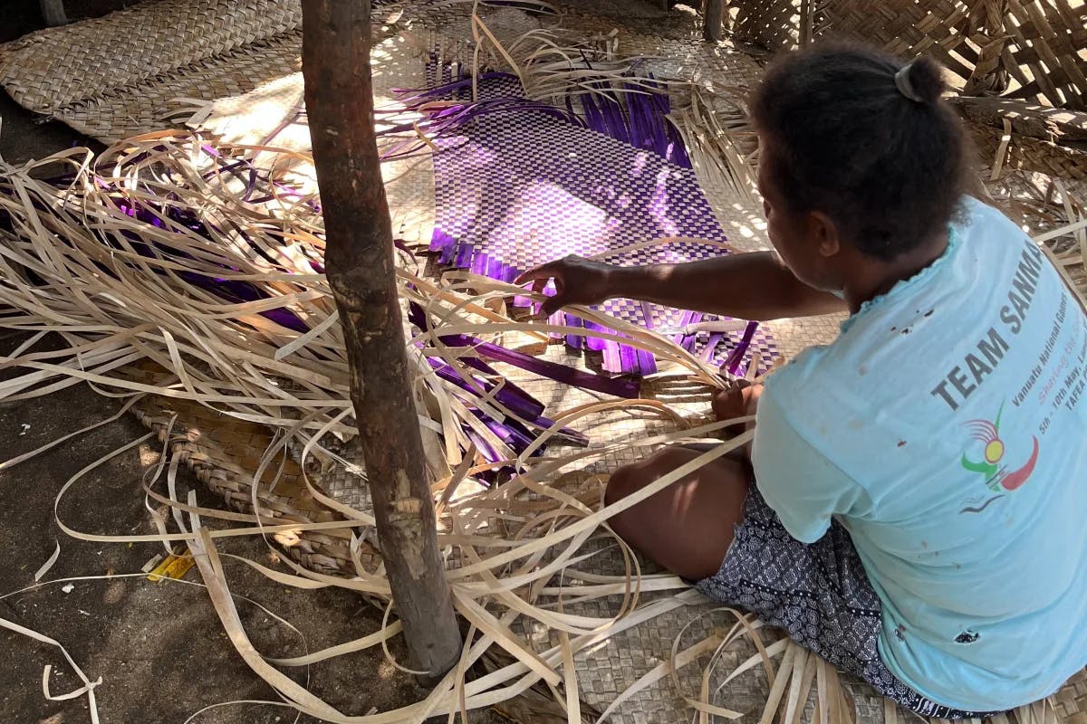 A person sitting on the ground weaving a purple and tan rug from straw.