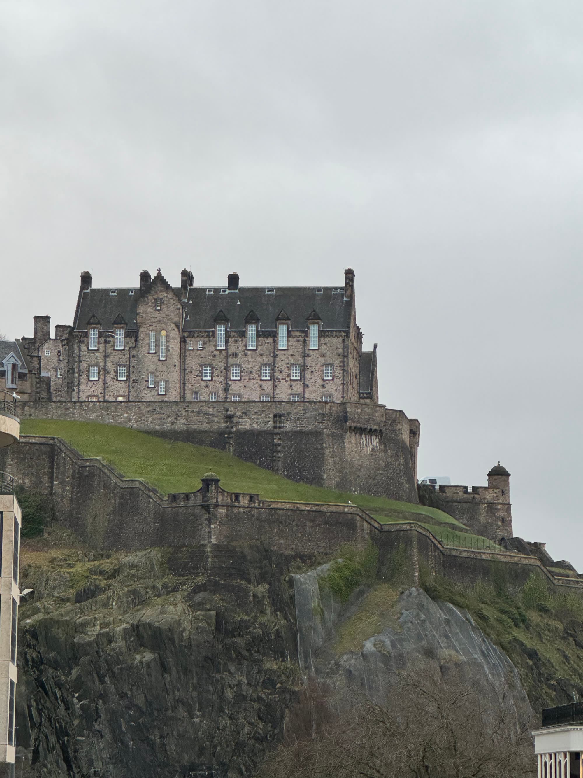 View of the Balmoral Edinburgh, a stone castle perched on a green cliff under cloudy skies